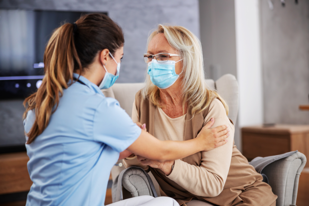 nurse-sitting-home-with-pensioner-comforting-her-during-coronavirus-pandemic-they-both-have-face-masks 1